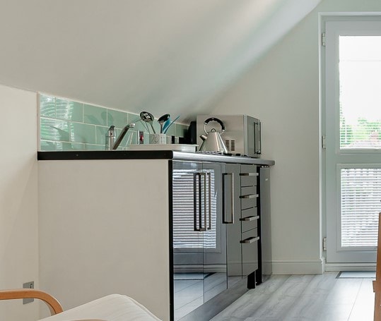 A compact kitchen features sleek black cabinetry with modern appliances, including a kettle and microwave. Natural light streams in through the large window with blinds, providing a bright atmosphere. Backsplash tiles in soft green add a subtle touch of color to the design.