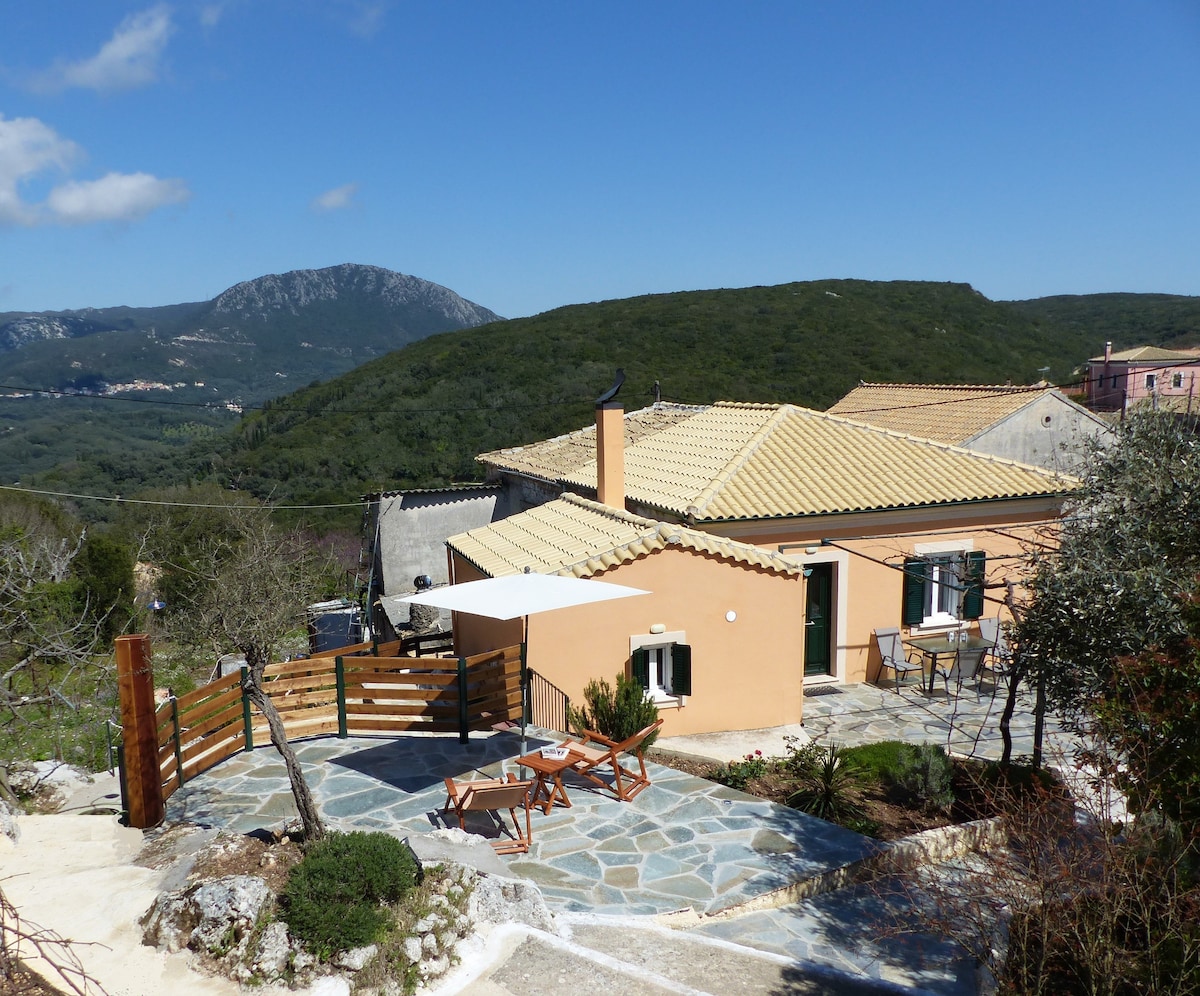 A modest house is seen surrounded by green hills, with a stone patio and outdoor seating area. A white canopy provides shade over a small table and chairs. A sunny sky fills the backdrop, enhancing the natural setting of the location.