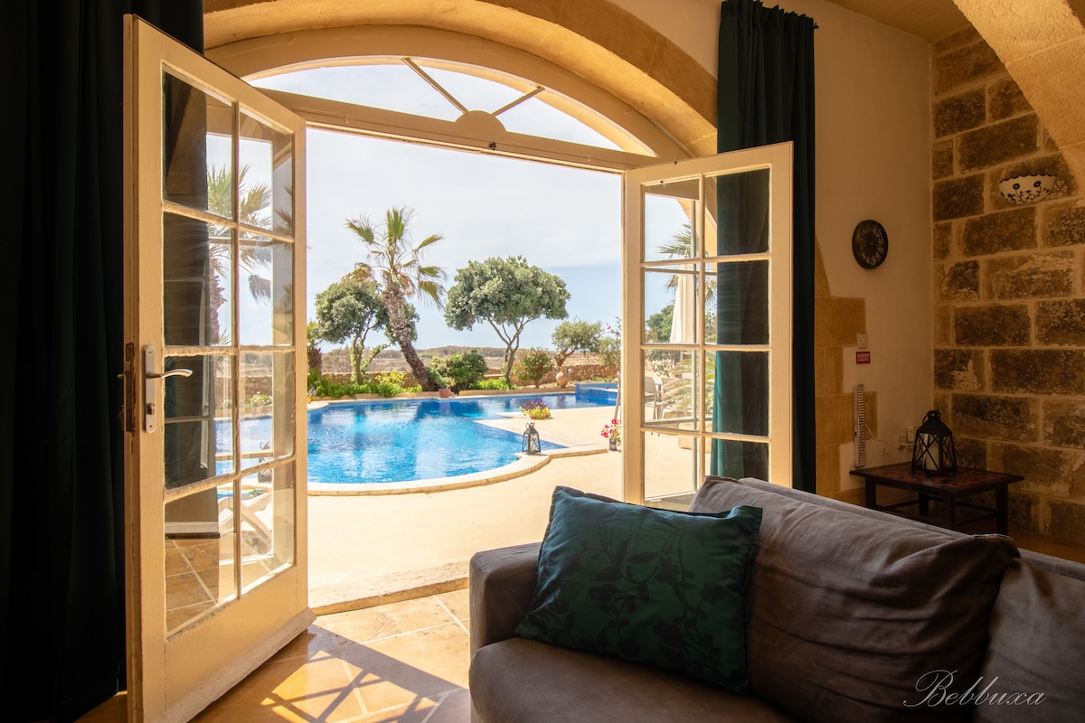 Double doors open to reveal a view of a large swimming pool surrounded by palm trees and lush greenery. Natural light floods the living space, where a cozy sofa is positioned, inviting relaxation and enjoyment of the serene outdoor setting.