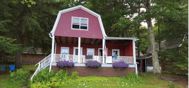 A charming cabin, painted in red, features a covered front porch with white railings and hanging flower baskets. Large windows allow for natural light, and the surrounding greenery enhances the connection to nature.