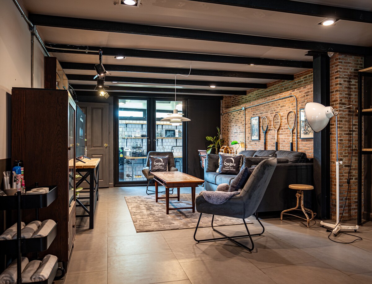 The living area features a mix of textured brick and dark wood, with large glass doors providing natural light. Comfortable seating includes an armchair and a sectional sofa, accompanied by a wooden coffee table atop a plush rug. Decorative artwork is displayed on the walls.