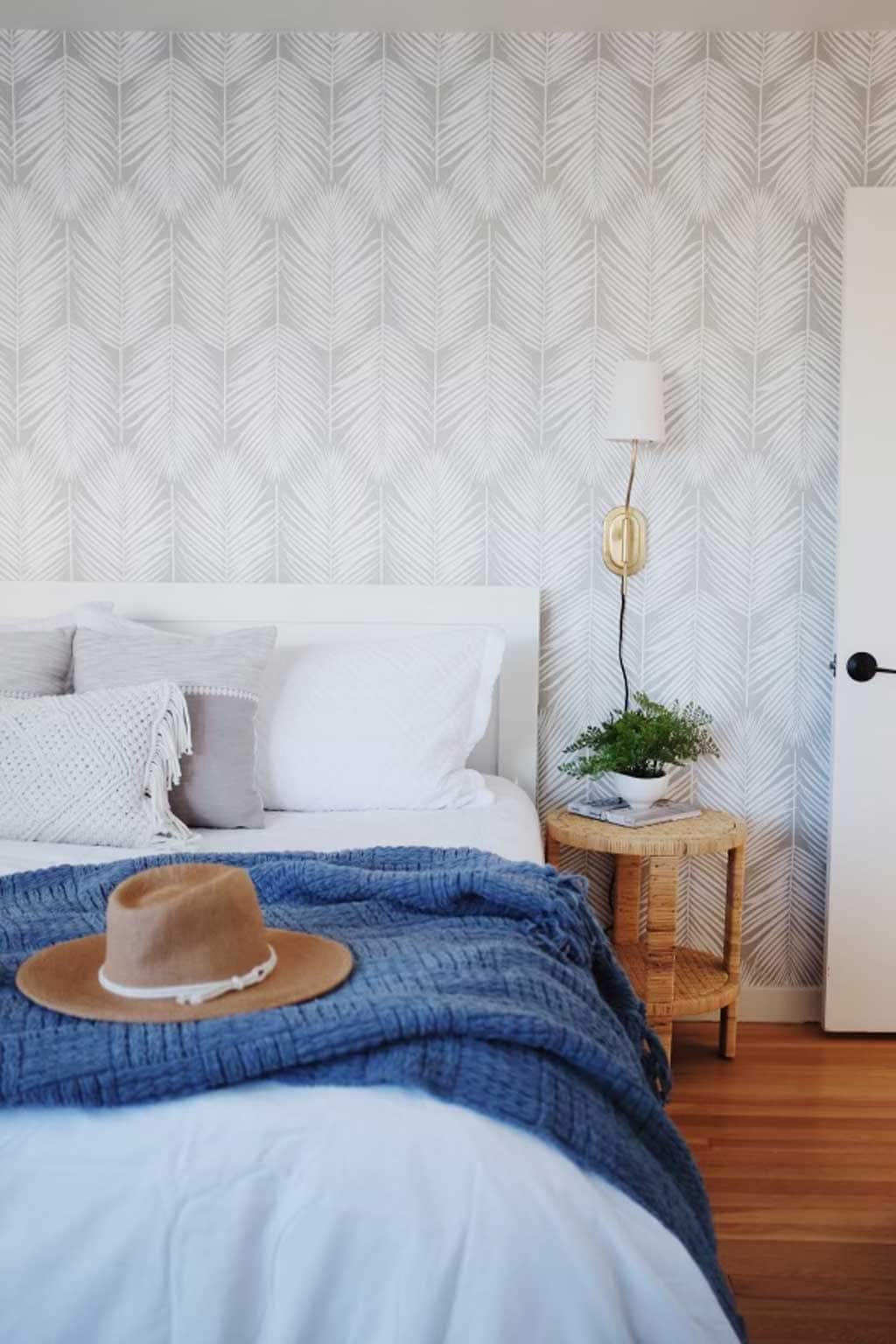 A bright bedroom features a spacious bed with white linens, complemented by a blue knitted throw and decorative pillows. A side table holds a small plant and a lamp, while a straw hat rests on the bed. The patterned wallpaper adds a subtle texture.