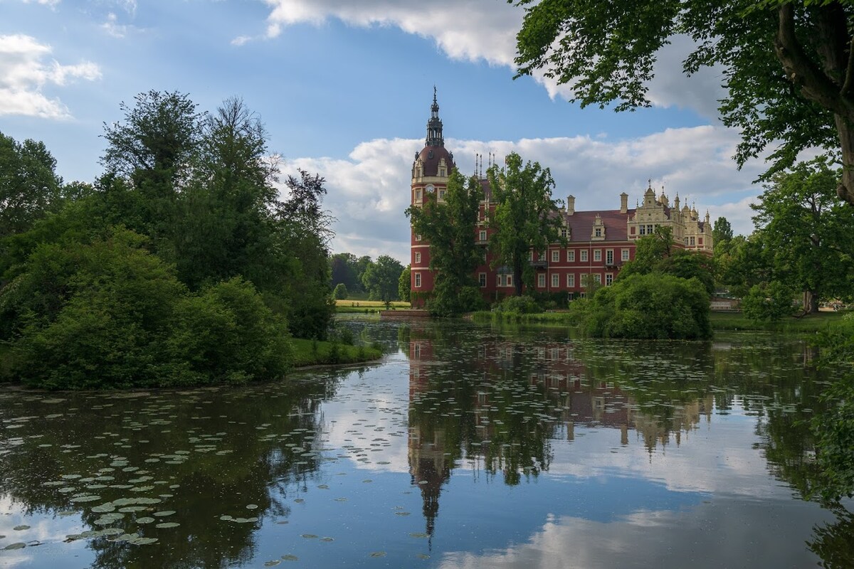 A historic building is reflected in tranquil waters, surrounded by lush greenery and trees. The structure features distinct architectural details and a tower, contributing to the picturesque scene. Floating lily pads can be seen on the surface of the water, enhancing the natural beauty of the location.