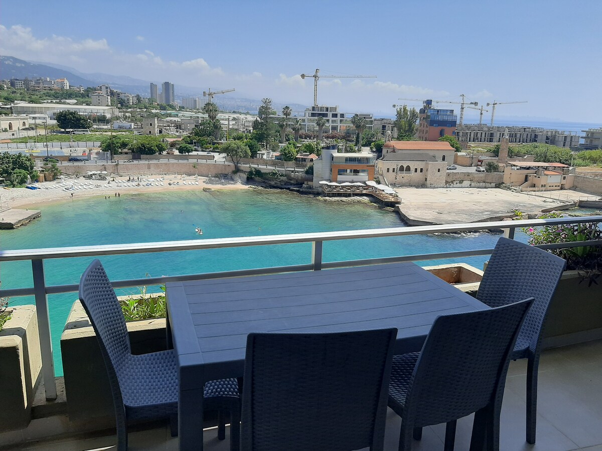 A terrace with a dining table and chairs overlooks a calm bay, featuring clear turquoise water and a sandy beach. A distant view of city buildings and cranes is visible against a bright blue sky.