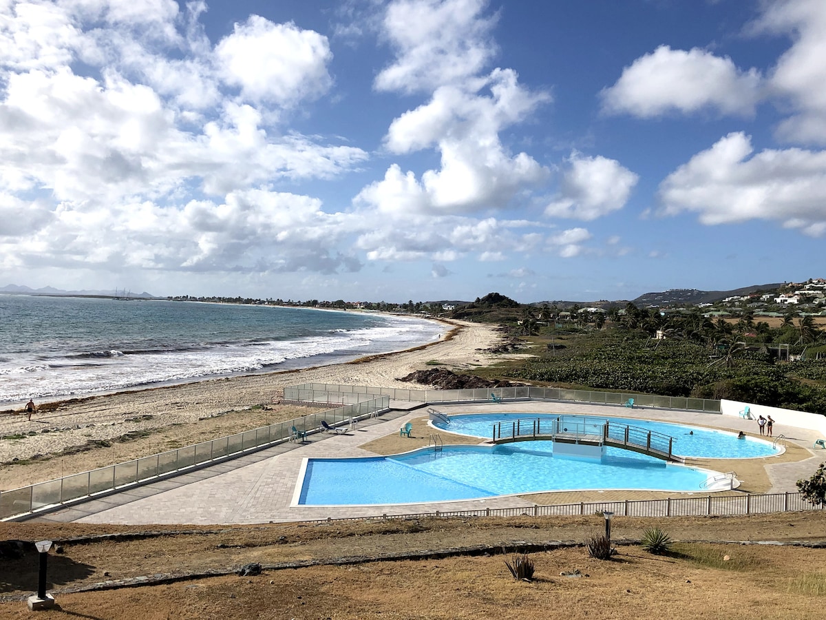 A spacious outdoor area features two swimming pools with clear blue water, surrounded by a patio and grass. The sandy beach and calm sea are visible in the background, with a picturesque shoreline under partly cloudy skies.