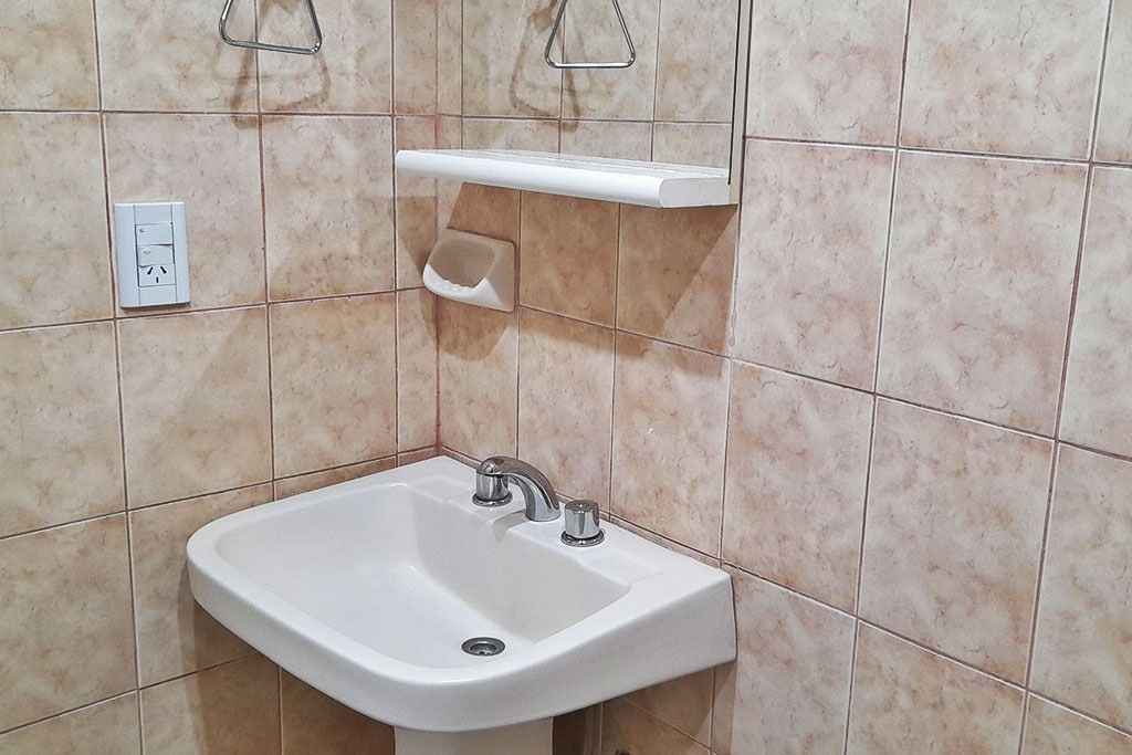 A small bathroom area is shown featuring a white sink with two faucets. The walls are tiled with light brown and beige tiles, creating a neutral backdrop. A mirror is mounted above the sink with a storage shelf beneath it and two towel hooks on the wall.