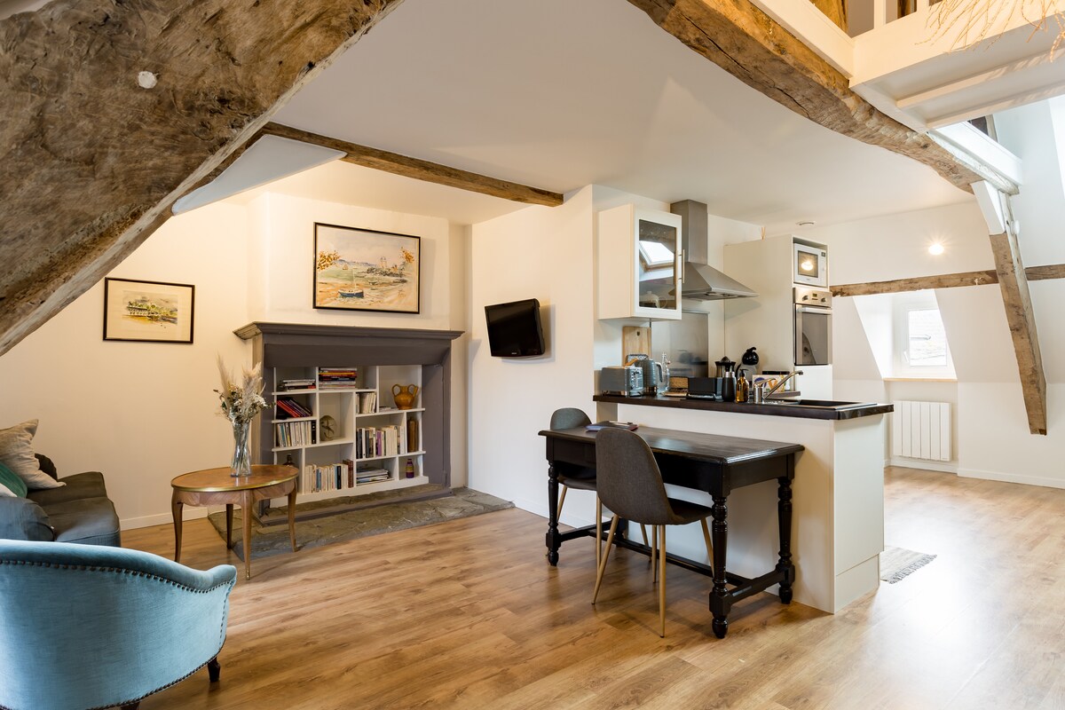 The image displays a spacious living area featuring old wooden beams and modern decor. A well-equipped kitchen is visible, along with a dining area that includes a table and chairs. A comfortable seating area with a couch is positioned nearby, enhancing a cozy ambiance.