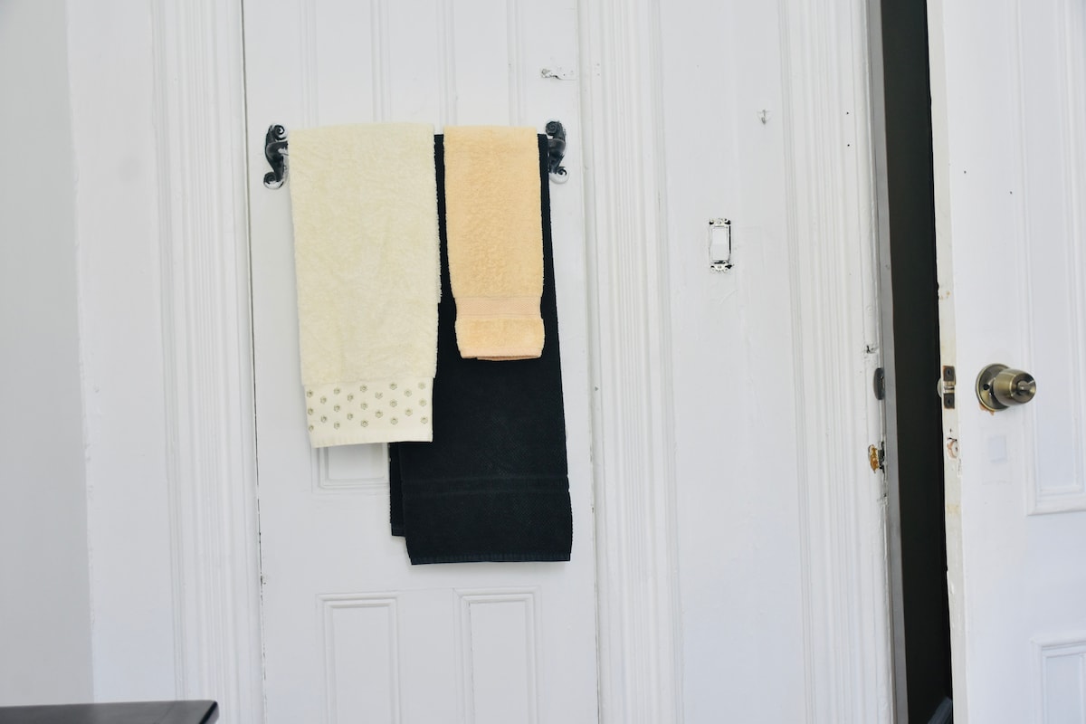 Two towels are hung on a wall-mounted rack next to a door. A cream-colored towel with a patterned border is positioned above a dark towel, providing essential bathroom amenities for guests. The doorframe and wall behind are painted in a clean white.