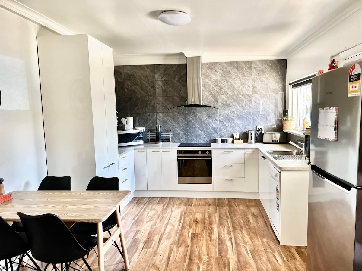 A modern kitchen features sleek white cabinets and a dark grey stone backsplash. A stainless steel refrigerator stands beside a spacious sink and counter. A dining table with black chairs is positioned in the foreground, enhancing the room's functional layout.