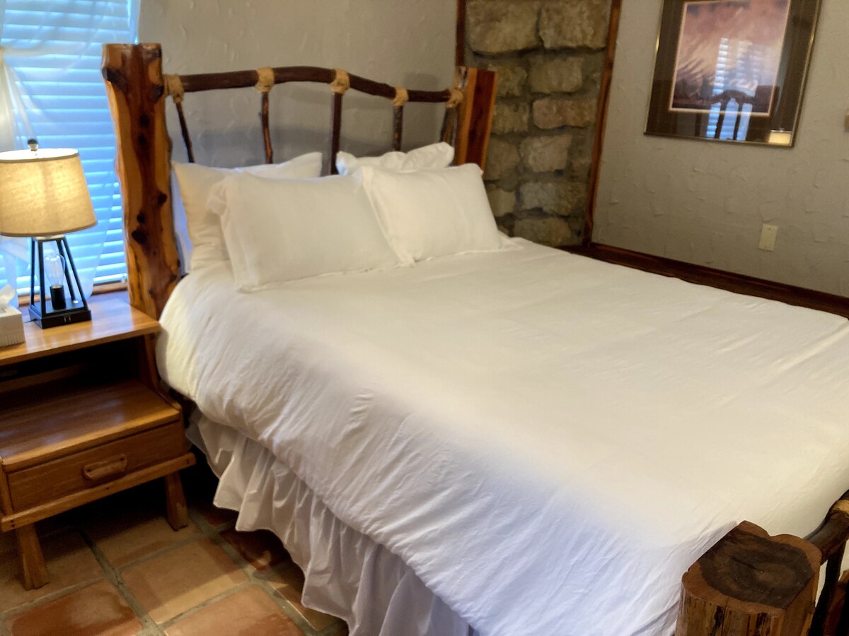 A cozy bedroom features a spacious bed with white linens and decorative ruffles. A bedside table with a lamp sits next to the bed, and a stone wall provides a rustic backdrop. Soft light filters through a nearby window.