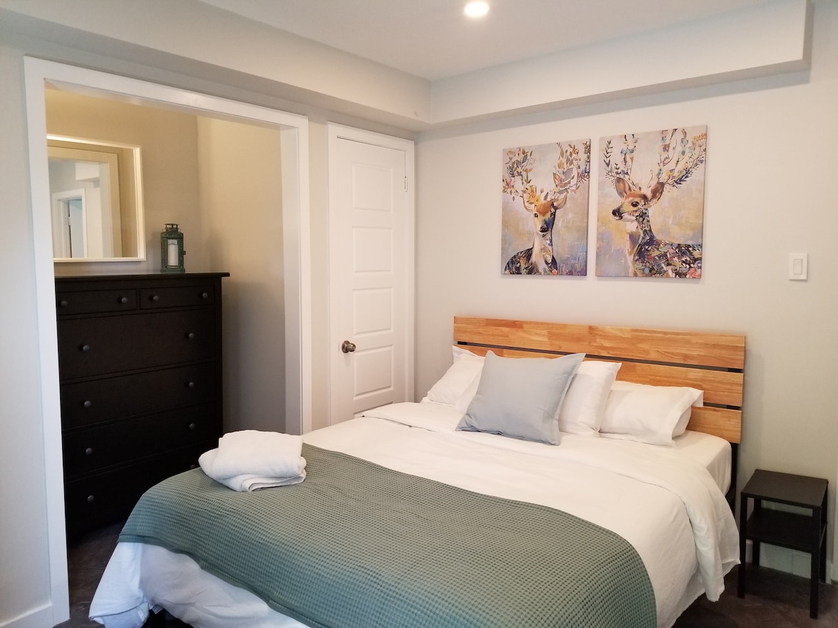 A cozy bedroom features a spacious bed layered with white linens and a green throw blanket. Two artistic deer paintings adorn the wall above the bed. A dark dresser is positioned nearby, and natural light is provided by a nearby mirror reflecting the room's calm ambiance.