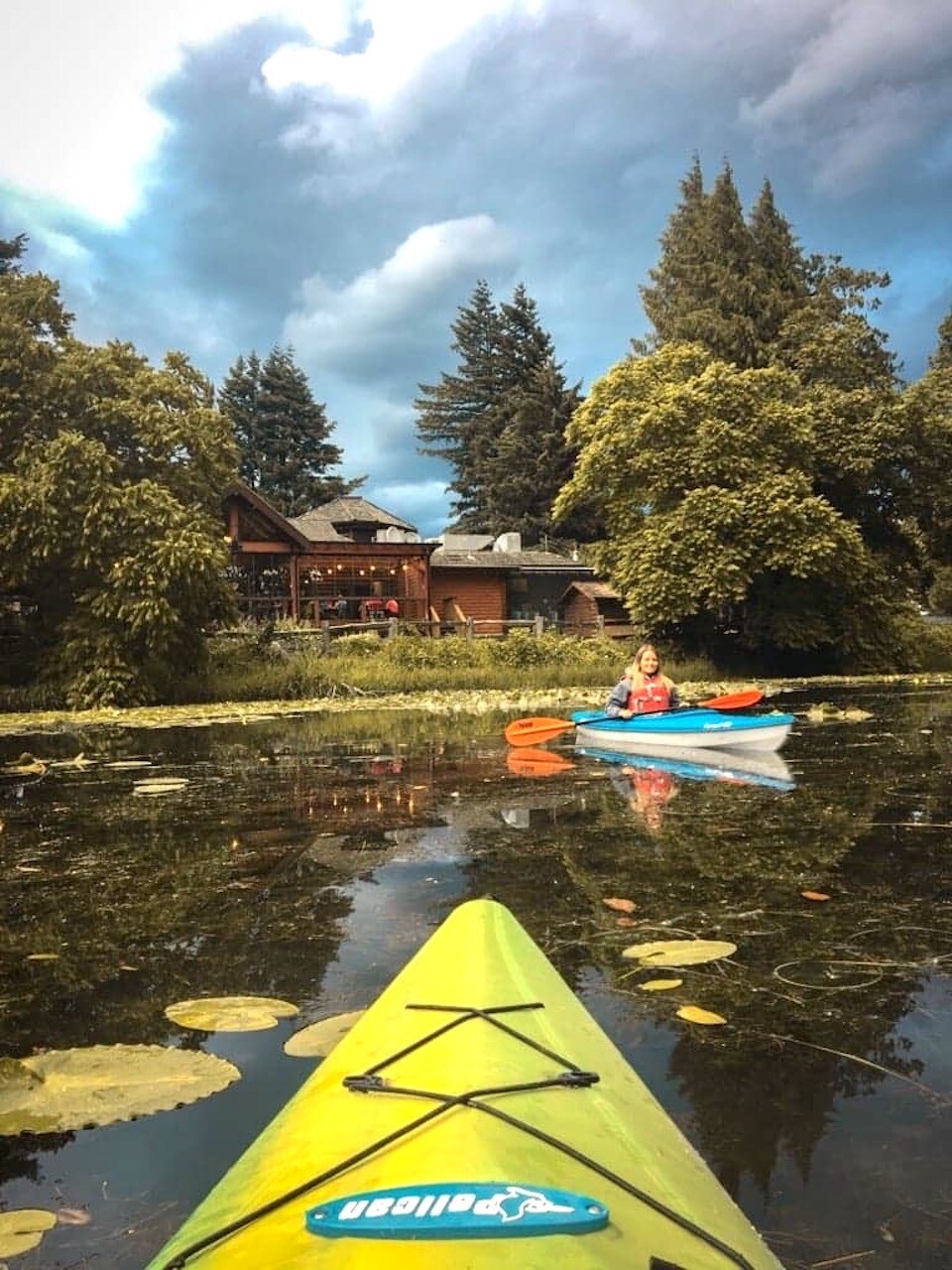 Two kayaks glide across a calm body of water covered in lily pads, with lush greenery framing the scene. In the background, a cottage is nestled among the trees, under a dramatic sky.
