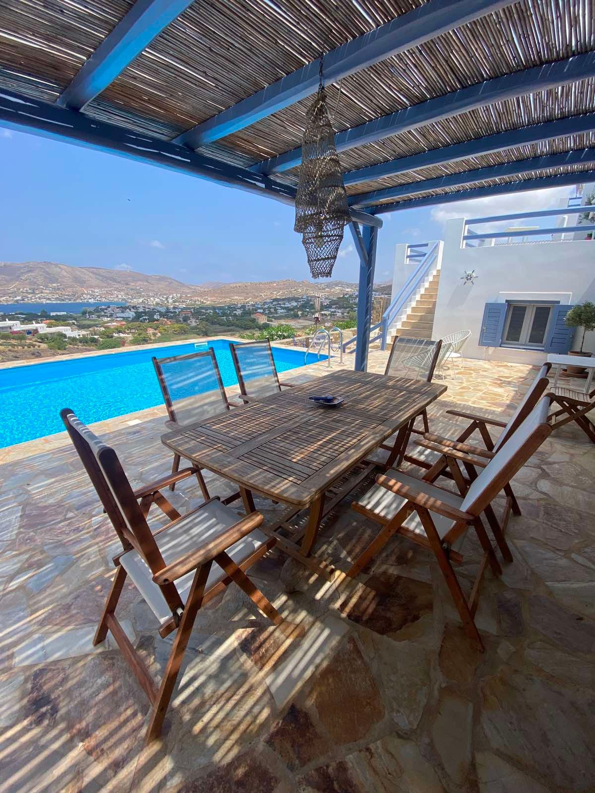 A shaded dining area featuring a long wooden table accompanied by several wooden chairs. The space overlooks a large swimming pool, with sweeping views of the village and surrounding hills visible in the background.