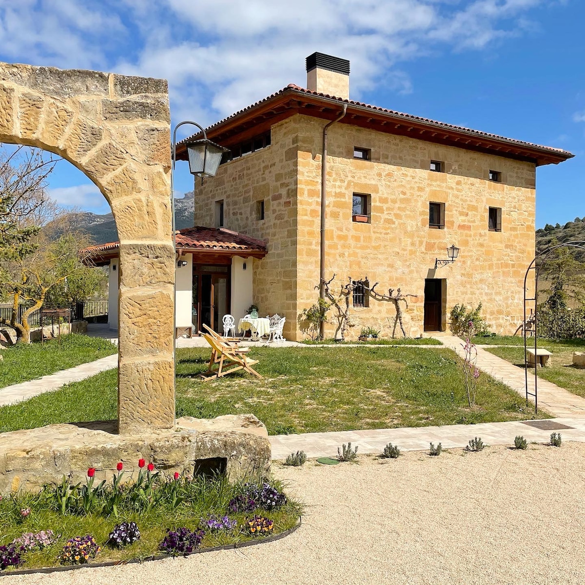 A recently renovated historic house is highlighted, showcasing its natural stone façade and warm red-tiled roof. Surrounding gardens feature flowers and greenery. A pathway leads to the entry, while a charming sitting area is visible, complete with outdoor furniture, offering a welcoming outdoor space.