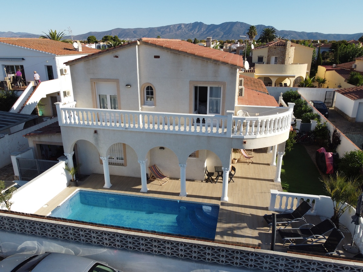 A villa featuring a spacious terrace with arched columns overlooks a private pool. The pool, located centrally, is complemented by sun loungers. A picturesque mountain backdrop is visible in the distance, enhancing the serene setting.