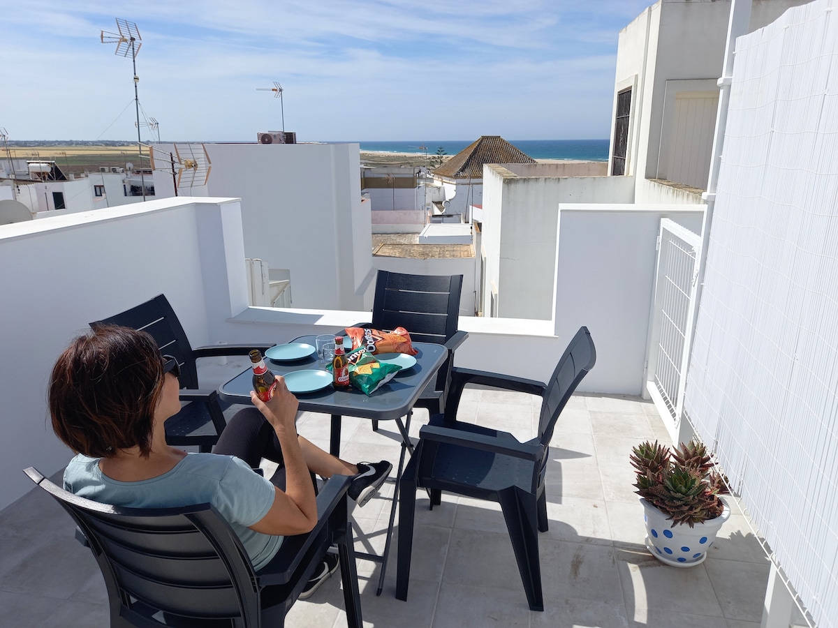 A terrace area offers a dining table set with chairs, accompanied by a colorful assortment of food and drinks. In the background, the ocean view extends beyond the white-walled buildings, under a clear blue sky. The setting provides an inviting space for outdoor relaxation.