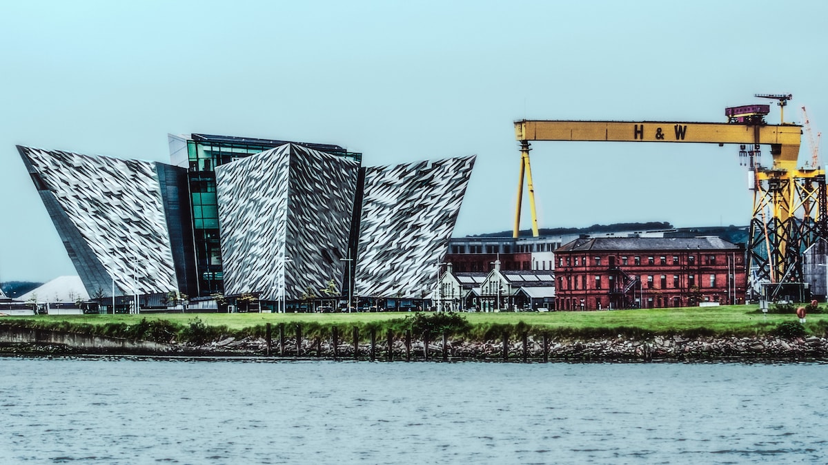 The image captures the Titanic Belfast visitor center, characterized by its striking, angular architecture that mimics the ship's hull. The Harland & Wolff cranes are visible in the background, offering a sense of industrial heritage alongside the waterfront.