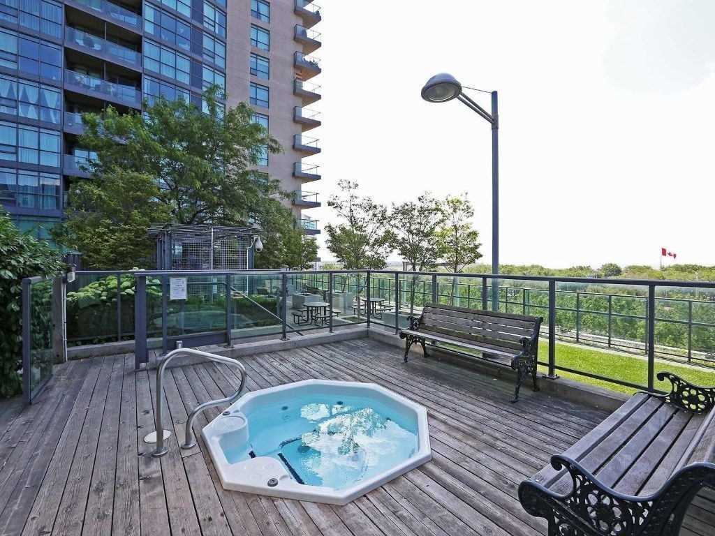 An outdoor area features a hot tub set within a wooden deck surrounded by greenery. Benches are positioned nearby, offering a place to relax. The skyline is visible in the background along with a flag fluttering gently, enhancing the serene environment.