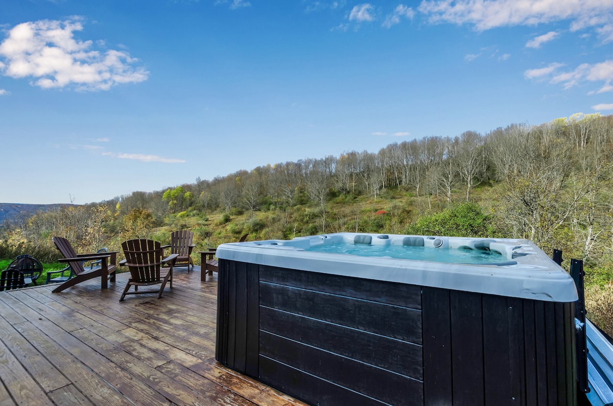 A private hot tub is positioned on a wooden deck, surrounded by Adirondack chairs. The scenic view features gently rolling hills and trees, complemented by a clear blue sky with soft clouds above.