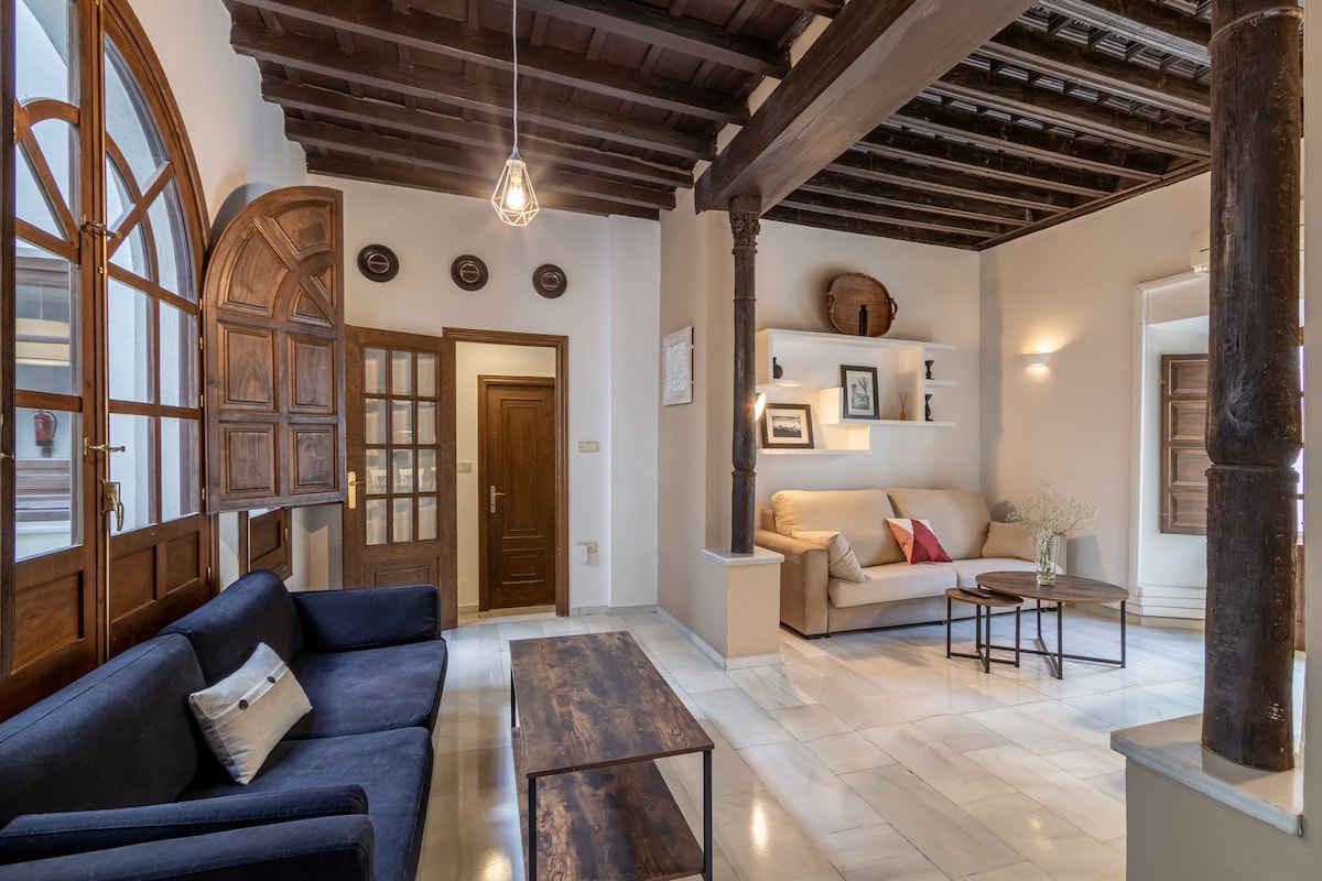 A spacious living area features high wooden ceilings supported by decorative pillars. Natural light fills the room through arched windows, illuminating a dark blue sofa and a light-colored couch. A coffee table centers the space, complemented by minimalistic wall shelves.