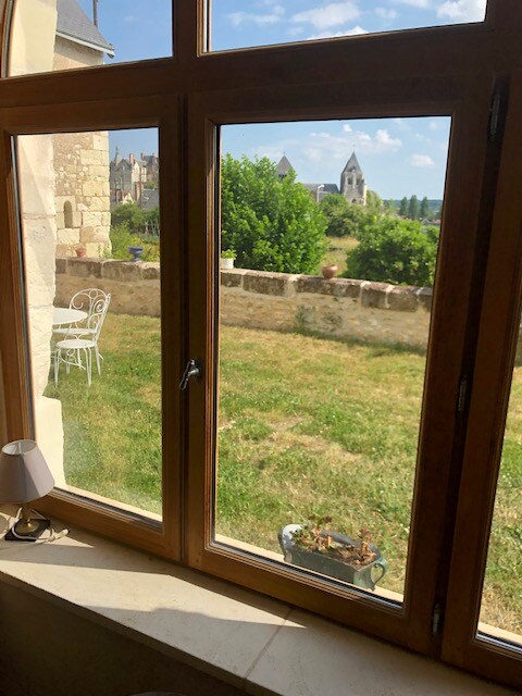 A wooden-framed window offers a view of a verdant garden and a distant historical building. Sunlight illuminates the outdoor seating area with a small table and chairs. The landscape is grassy and well-kept, enhancing the connection to nature.