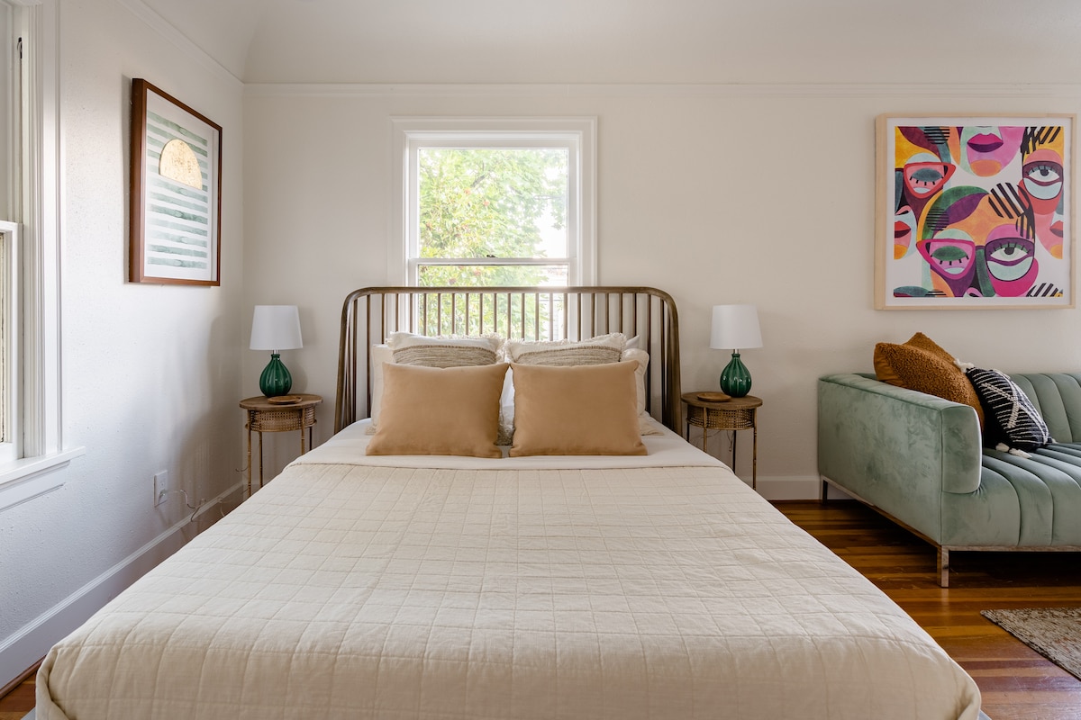 A bright sleeping area features a spacious bed with a neutral-colored quilt, accented by two plush pillows. Soft light filters through a window, illuminating the room. A comfortable green sofa and two bedside tables with lamps create a cozy atmosphere.