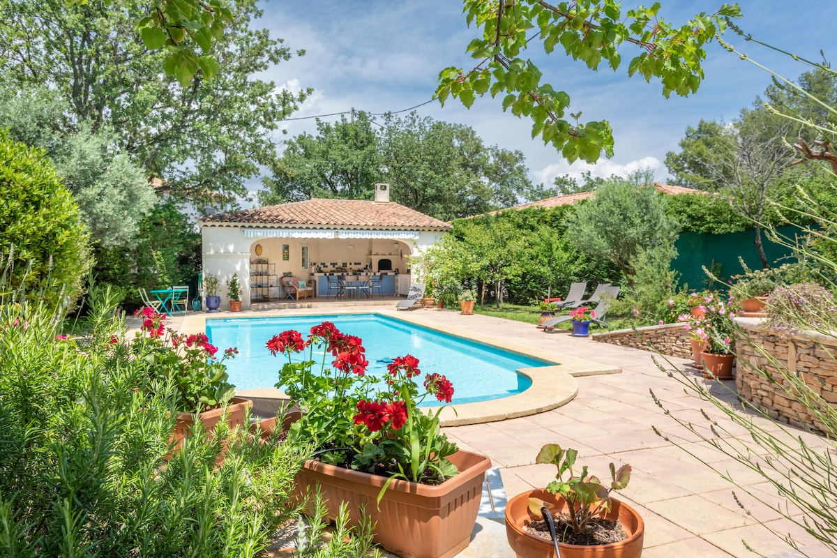 A serene outdoor area showcases a sparkling swimming pool surrounded by lush greenery and vibrant flowers in terracotta pots. A covered patio with seating overlooks the pool, providing a perfect space for relaxation and dining in nature.