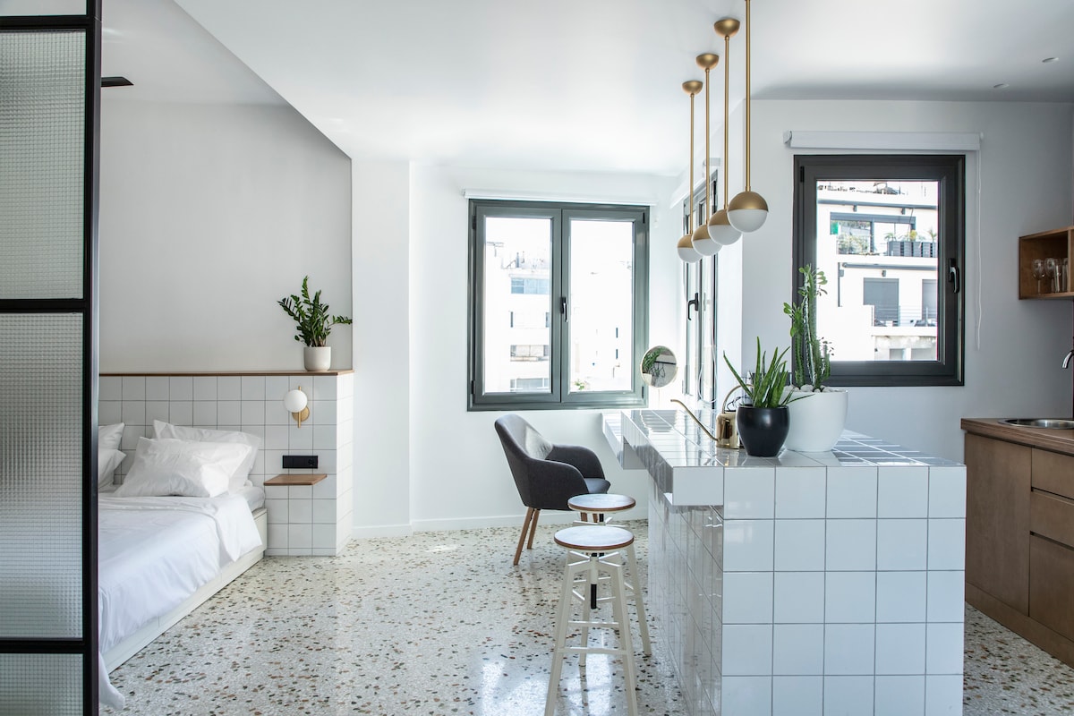 An open-plan living space features a cozy sleeping area with a spacious bed, complemented by modern furnishings. A small dining table with two stools is positioned near a window, while the kitchen area showcases sleek cabinetry and a minimalist design. Natural light filters in through large windows.