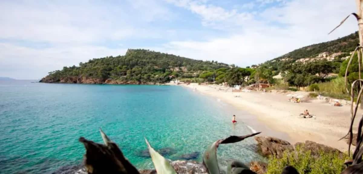 A tranquil beach scene features gentle waves lapping against the sandy shore. Lush green hills line the background, dotted with trees. The clear turquoise water is contrasted by a sandy beach where a few visitors can be seen enjoying the serene surroundings.