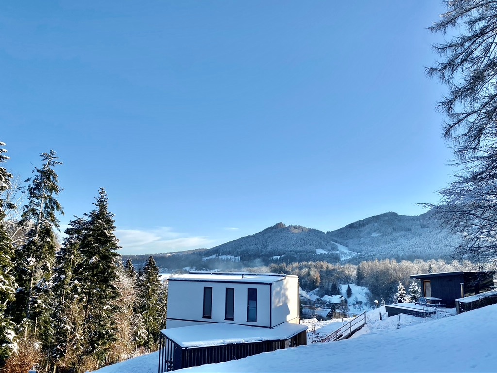 A modern building is visible in the foreground, partially covered in snow. Surrounding trees are dusted with white, and rolling hills are seen in the distance under a clear blue sky.