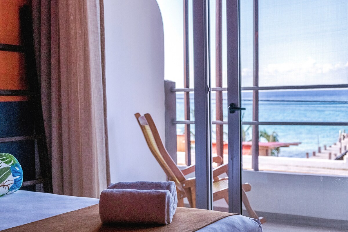 A serene view from an inviting room is showcased, featuring a spacious bed with a rolled towel placed neatly on a golden blanket. French doors open to a private balcony, revealing a glimpse of the ocean and wooden deck below.