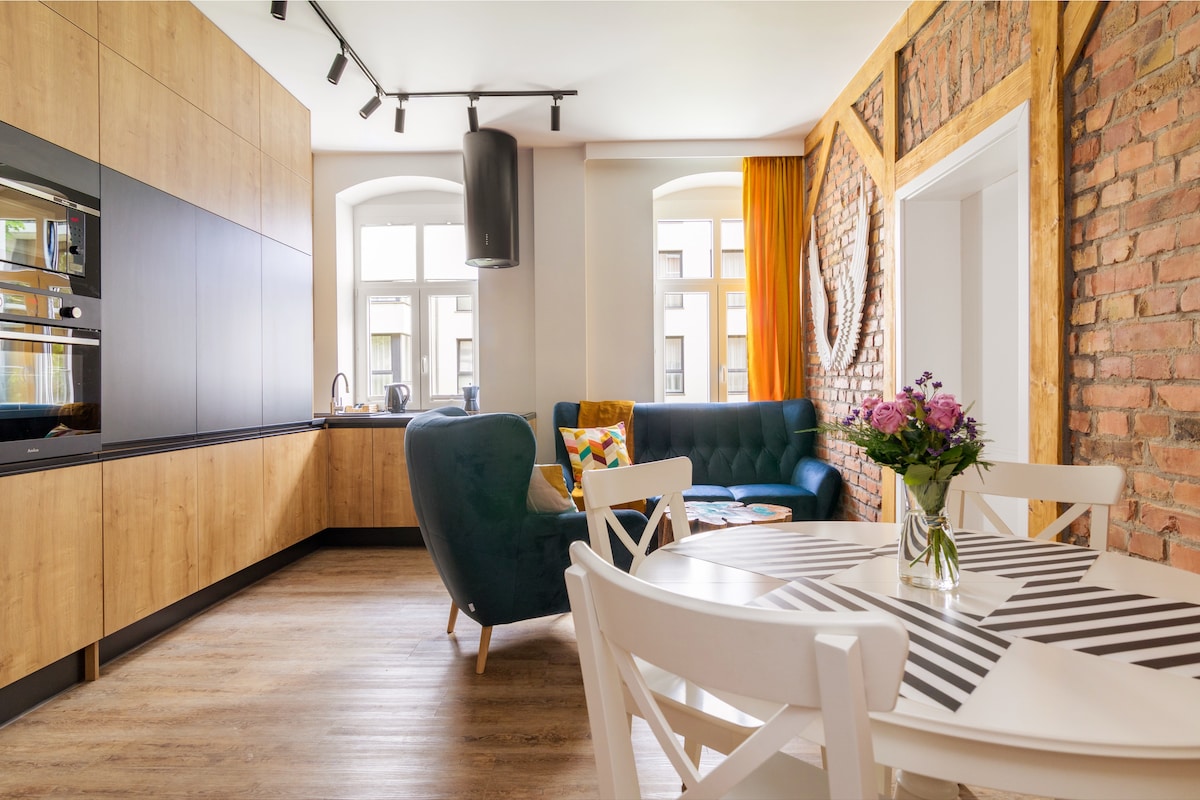A modern living area is presented with a teal sofa and a circular mirror against a textured brick wall. A round dining table with a striped pattern is set beside the kitchen, which features wooden cabinetry and built-in appliances. Natural light enters through the large windows.