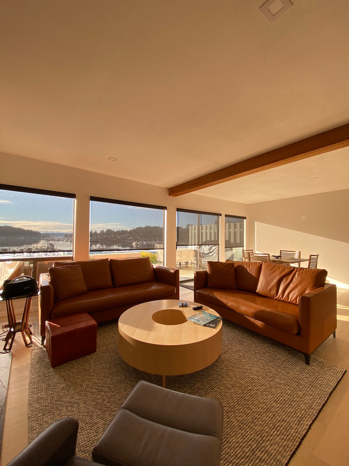 A modern living space features two brown leather sofas positioned around a circular wooden coffee table. Large windows provide expansive views of the marina and surrounding landscape, allowing natural light to fill the room. A textured area rug adds warmth to the wooden floor.