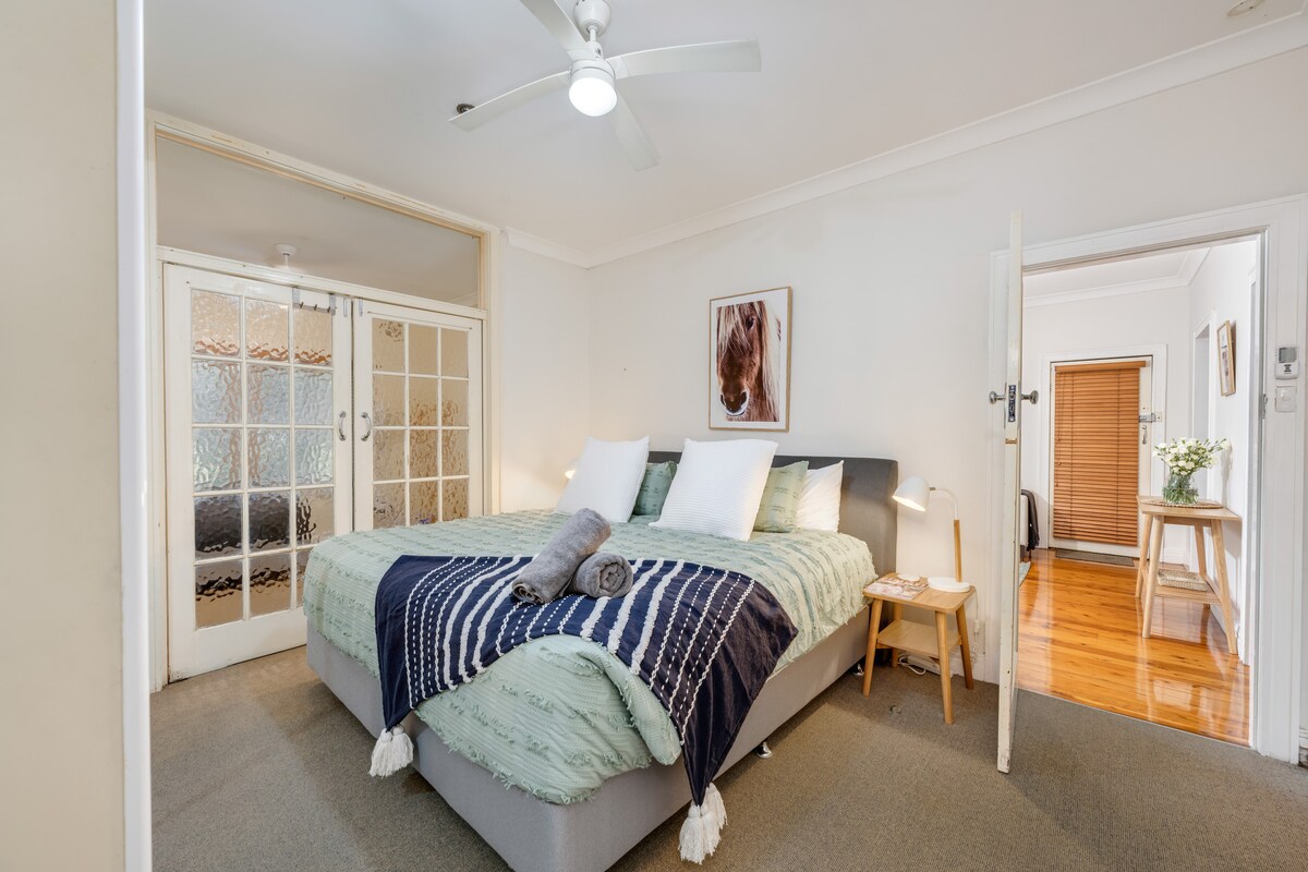 Posteľ Super King s kvalitnou posteľnou bielizňou, nadýchanými vankúšmi, stropným ventilátorom, zrkadlovým šatníkom. Dvere prechádzajú do jednolôžkovej spálne. 