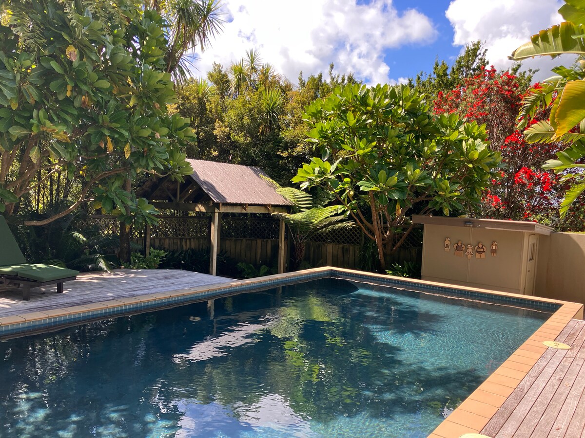 A secluded swimming pool area is surrounded by lush greenery and colorful flowering plants. A wooden deck offers space for lounging, and a shaded gazebo provides shelter. The pool, fully fenced, features clear blue water reflecting the sky and nearby foliage.
