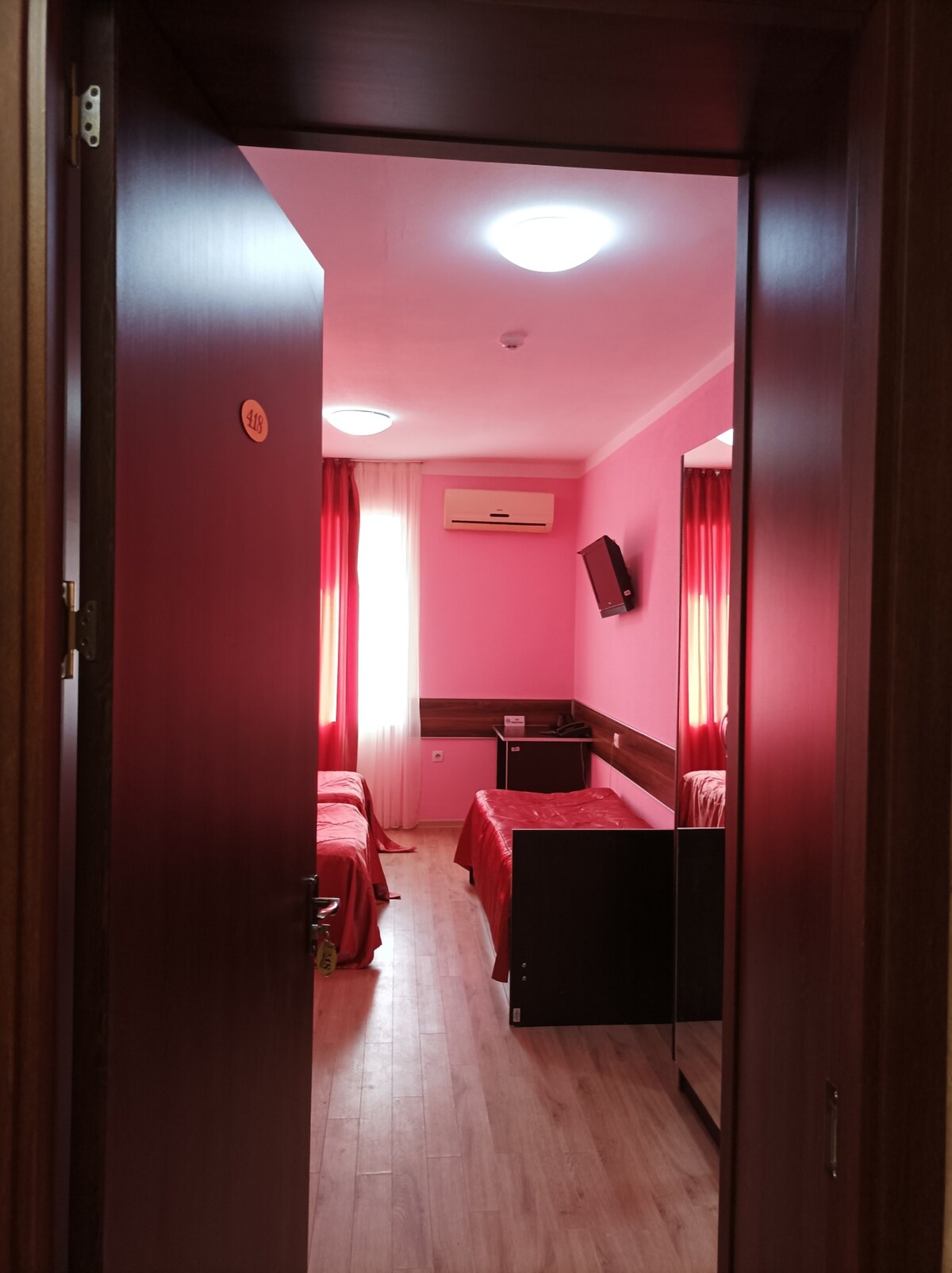 A hallway view leads into a room featuring two beds dressed in red linens. Pink walls and red curtains provide a warm ambiance. A television is mounted on the wall, with a nightstand positioned between the beds and a mirror visible to the side.
