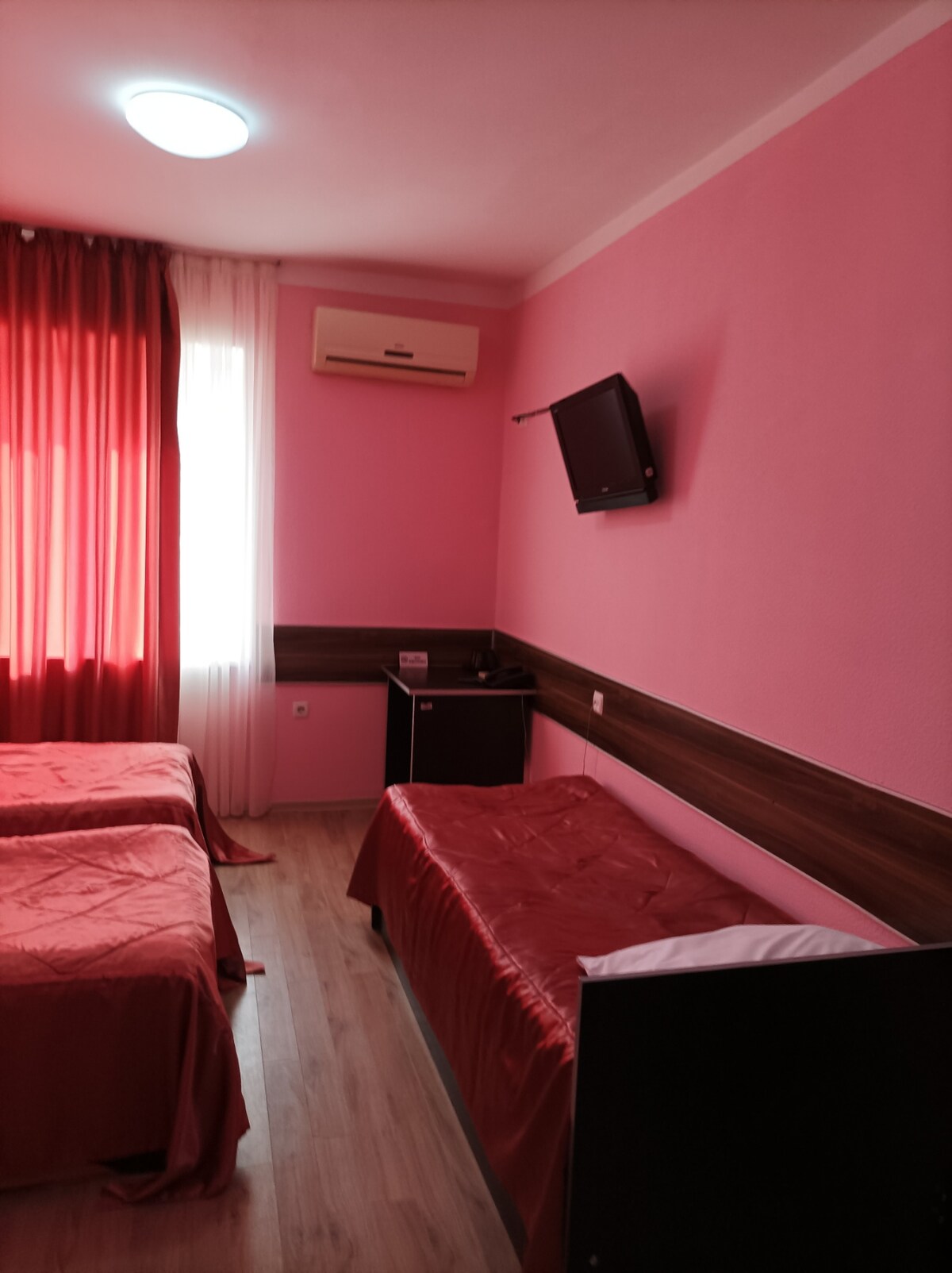 A bright room features two twin beds dressed in red coverlets. A wall-mounted television is positioned above a wooden dresser. Large windows with sheer and heavy red curtains allow natural light to fill the space, complemented by light-colored wooden flooring.