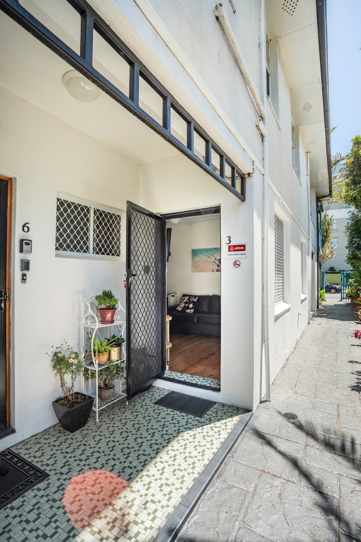 An entranceway to the ground floor unit is visible, featuring a screen door and potted plants. The indoor area reveals a cozy lounge space with a dark couch, complemented by a light-colored wall and artwork. Natural light brightens the entry, enhancing visibility.