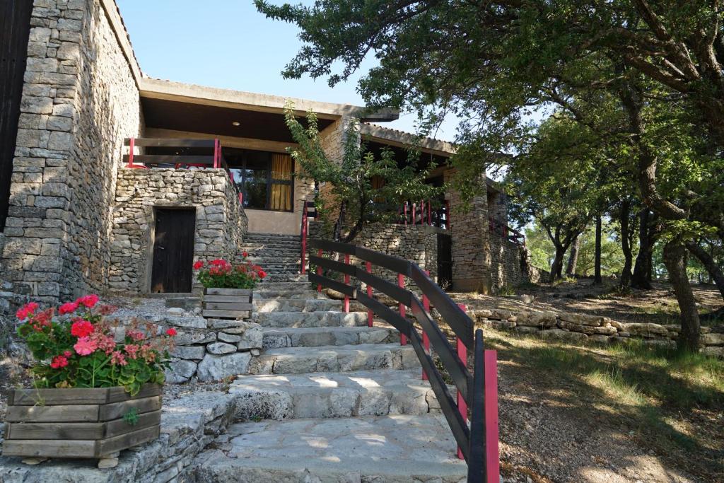 A stone-built residence is depicted, set against a natural landscape. Steps lead up to the entrance, flanked by vibrant flower planters. The structure features wooden railings and is shaded by large trees, creating a peaceful outdoor setting.