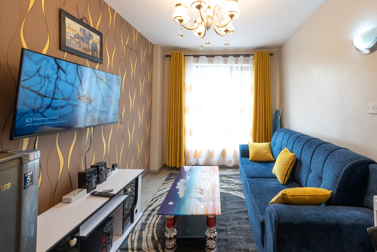 A modern living area features a blue upholstered sofa adorned with bright yellow cushions. A glass-top coffee table sits in front, and a large television is mounted on the wall. Floor-to-ceiling curtains adorn the window, allowing natural light to brighten the space.