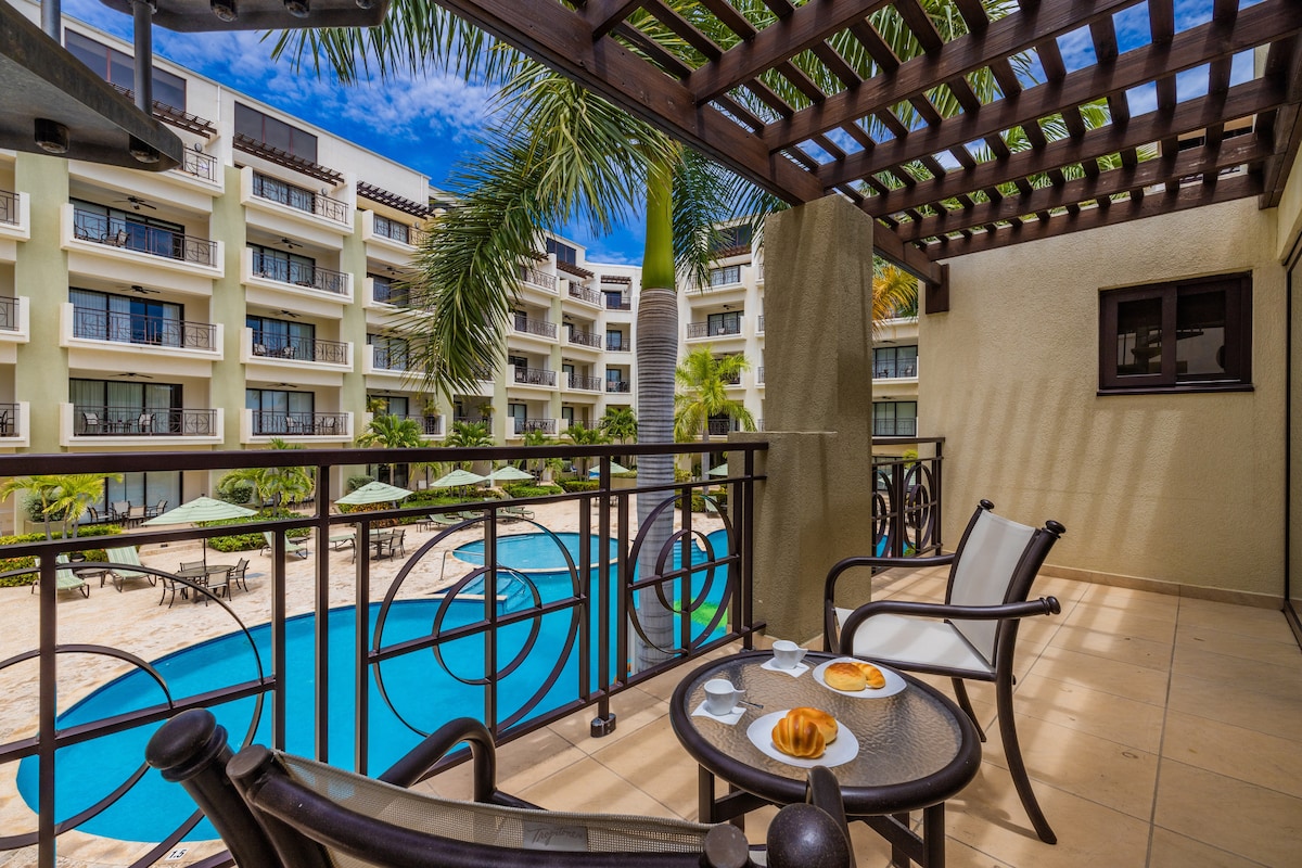 A cozy balcony is showcased with two chairs and a table, offering a view of the pool area surrounded by lush greenery. Light pastries are placed on the table, highlighting a tranquil space for relaxation.