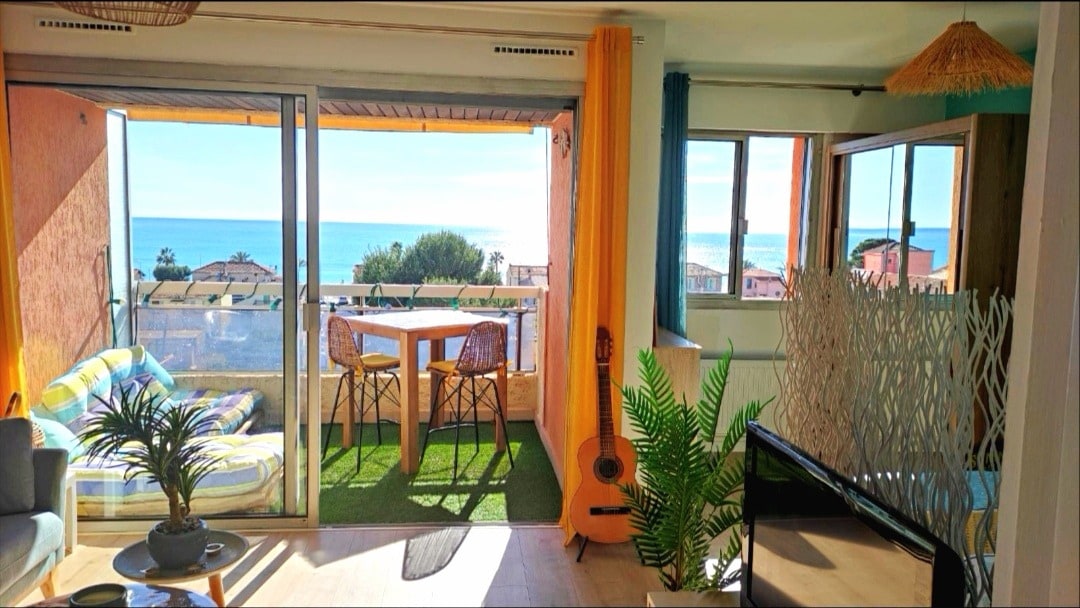 A bright living area is showcased with a large sliding glass door opening to a balcony with a sea view. A small wooden dining table sits on the balcony, while a cozy sofa and decorative plants create a warm atmosphere inside.