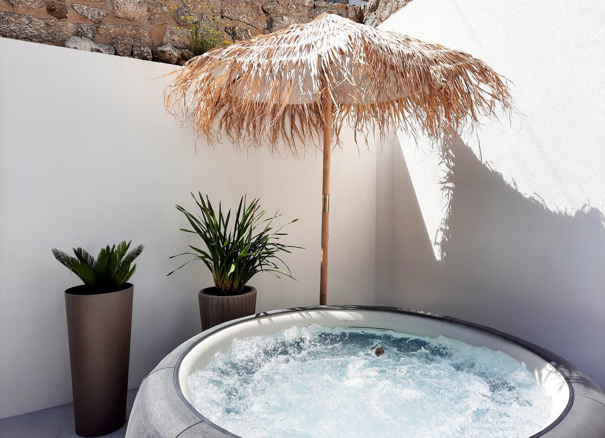 A private outdoor space features a round hot tub surrounded by minimalist white walls. A straw parasol provides shade next to two potted plants, adding greenery to the area. The clear water bubbles gently, creating a serene atmosphere.