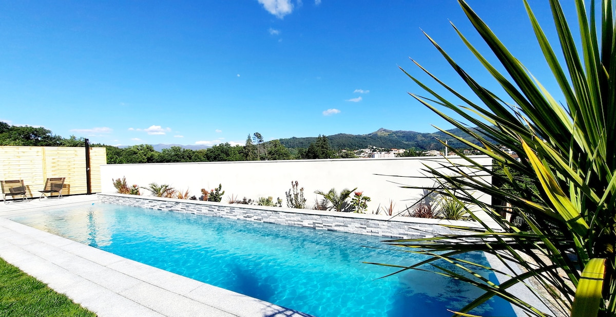 An outdoor swimming pool features clear blue water, bordered by a sleek stone deck. Lush greenery surrounds the pool area, with a few sun loungers positioned for relaxation. A panoramic view of distant hills is visible against the backdrop of a clear blue sky.