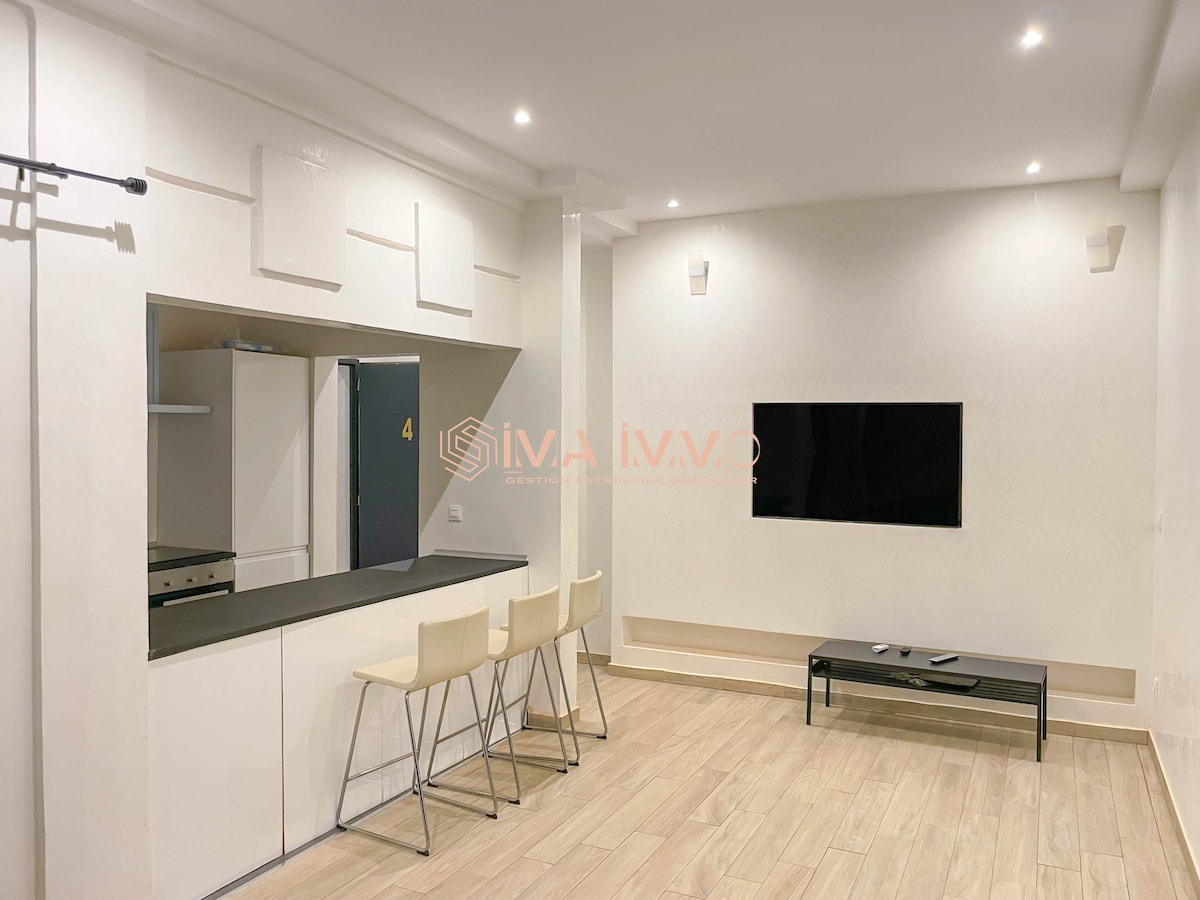An open living area features a sleek kitchen counter with four stools. A flat-screen television is mounted on the light-colored wall opposite, while a modern kitchen with built-in appliances is visible in the background. Soft lighting creates a bright and welcoming ambiance.