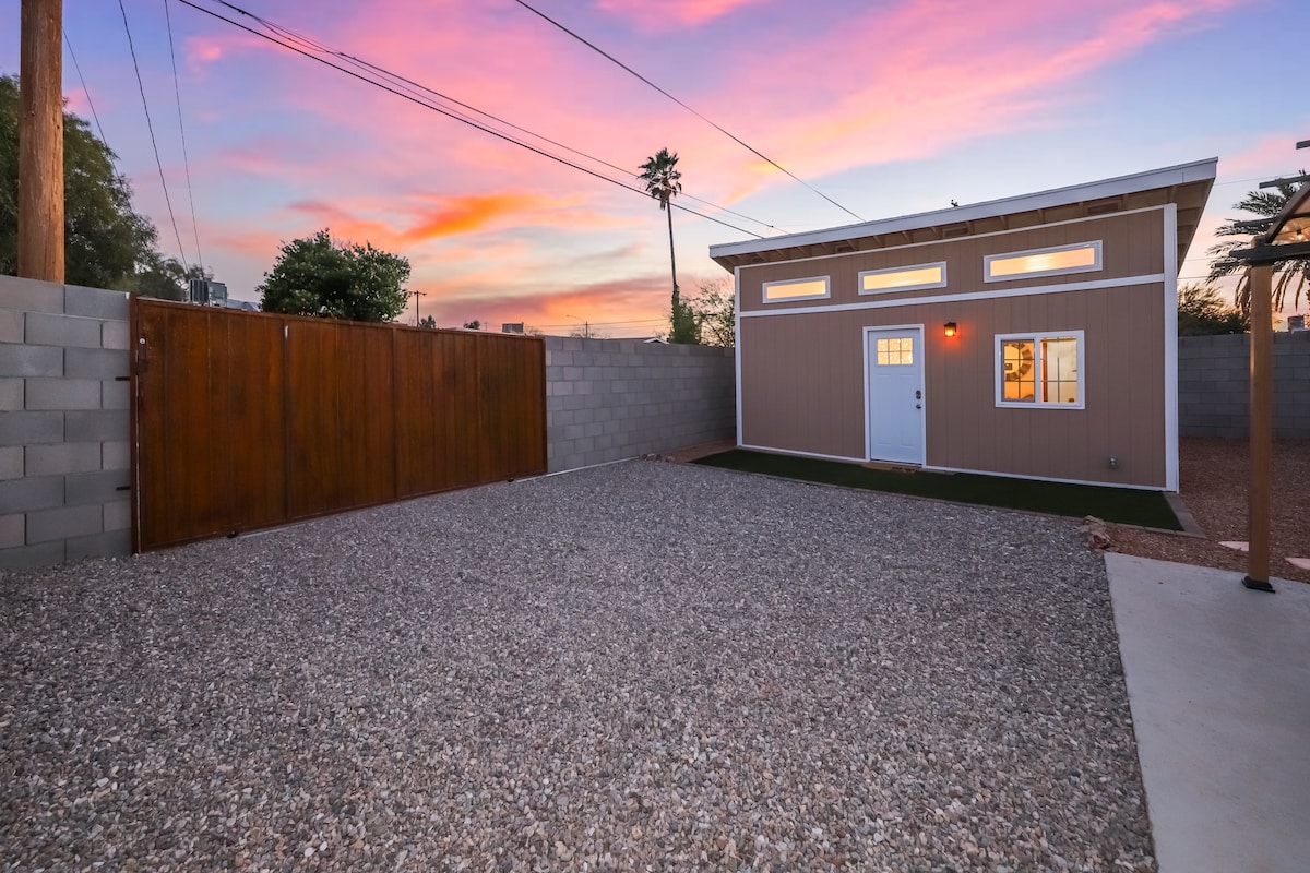 An exterior view of the Tiny Smart home is showcased during sunset, featuring a gravel parking space and a wooden gate. The home displays a welcoming entrance with large windows that offer a glimpse inside, while a colorful sky adds a serene backdrop.