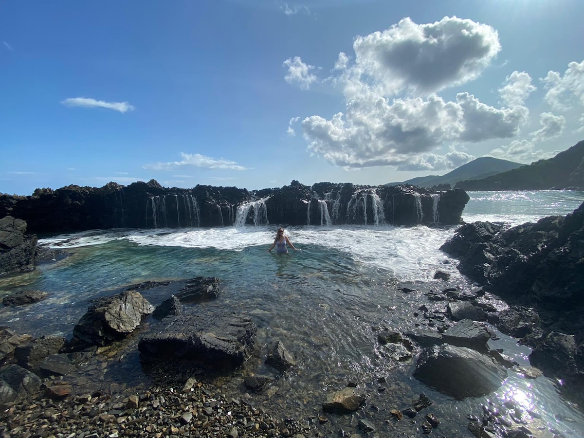 A person is seen enjoying a serene moment in a natural pool formed by rugged rocks along the shoreline. Gentle waves create a soft splash against the rocks, while the sky displays a mix of white clouds and blue hues, enhancing the tranquil setting.
