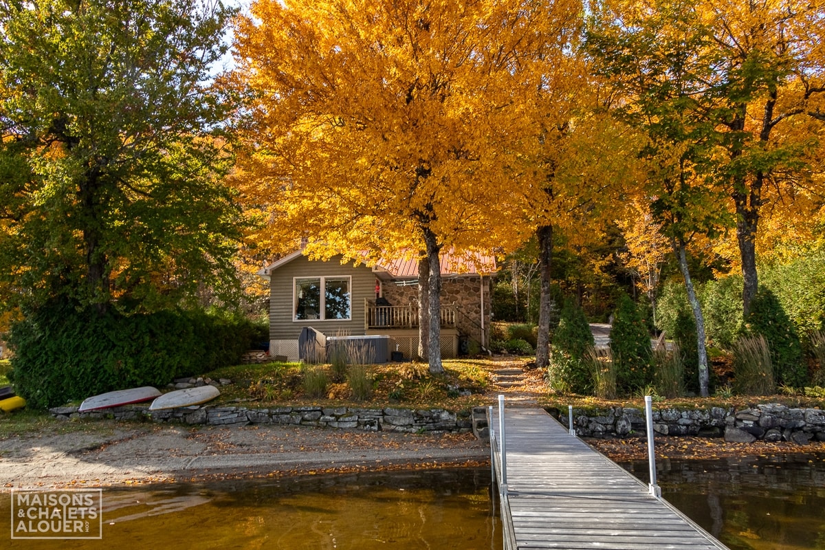 A cabin is situated near a sandy beach, surrounded by vibrant autumn foliage. A wooden dock extends into the calm waters, while paddleboards and kayaks rest along the shore. The serene setting is complemented by the clear blue sky.