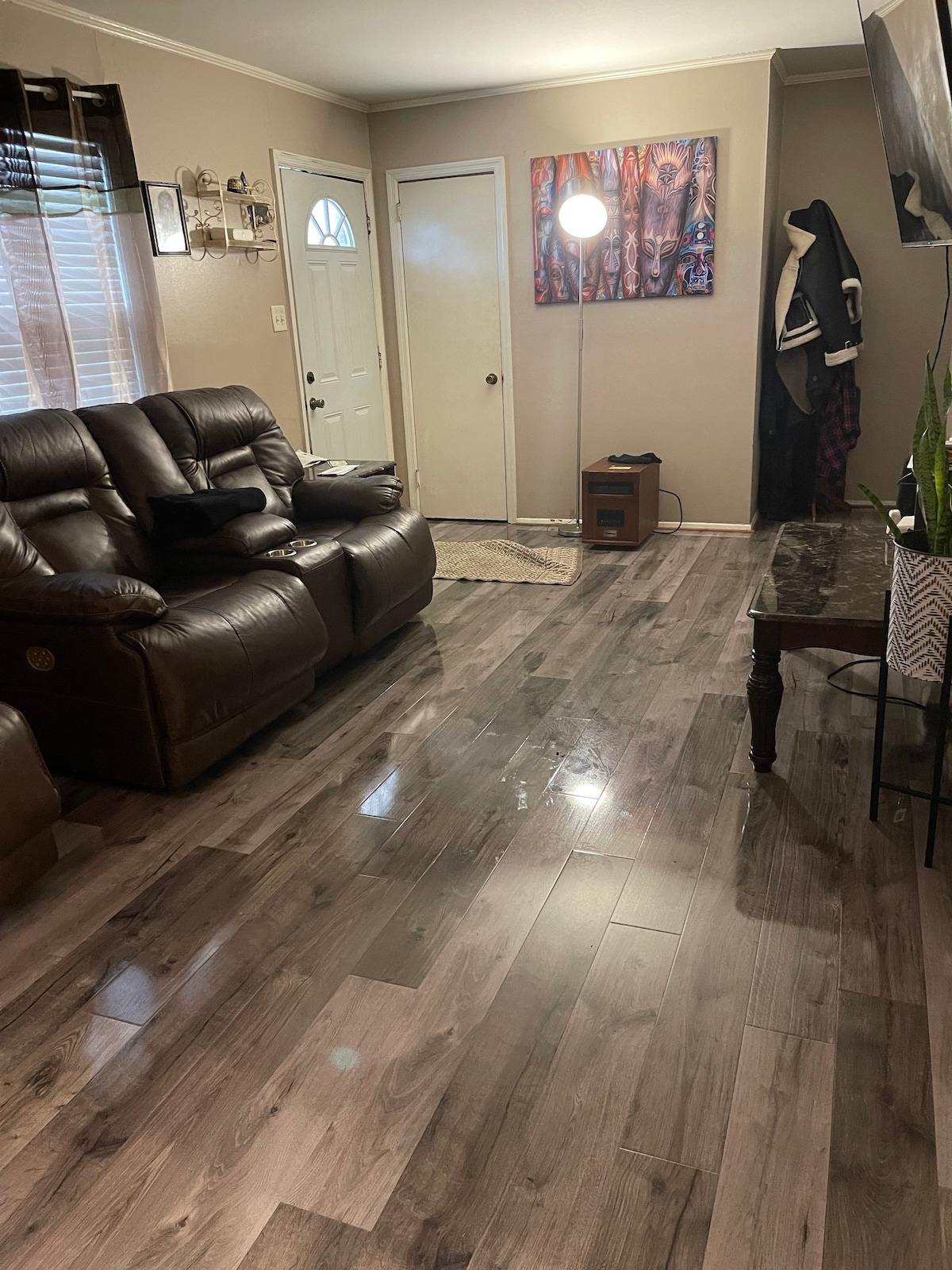 A living area features dark leather seating arranged for comfort, with a floor lamp providing soft illumination. Light reflects off the polished wooden floor, which is complemented by a textured area rug. A wall-mounted TV is visible, alongside a decorative wall art piece.