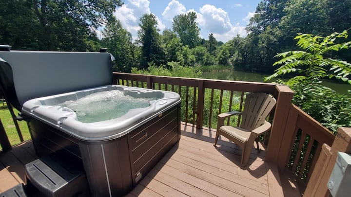A private hot tub is situated on a wooden deck, overlooking a serene pond surrounded by lush greenery. A single chair is placed nearby for relaxation, while the sun casts a bright glow on the peaceful natural setting.